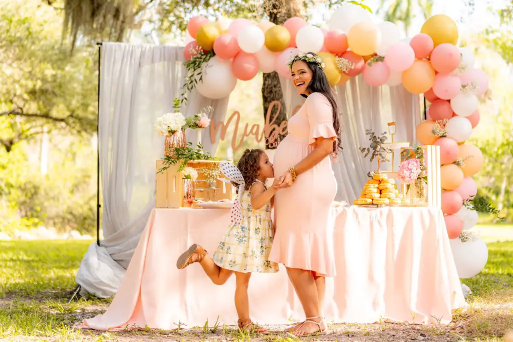 Little girl kissing her pregnant mother’s belly in front of baby shower decorations. - How To Plan A Baby Shower Checklist - Simple Guide to Planning A Brilliant Baby Shower - Baby Journey blog