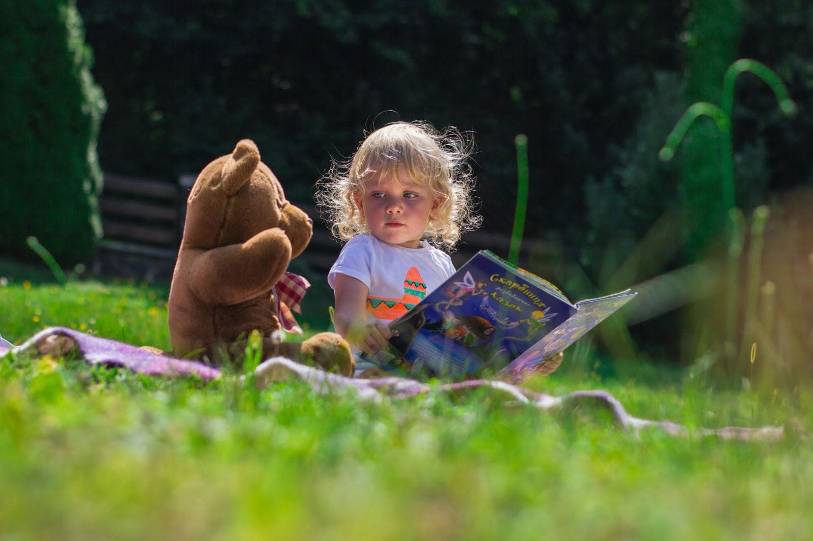Cute girl toddler sitting on the grass holding a big picture book while looking at her bear soft toy - Baby Names That Mean Curious - Baby Journey