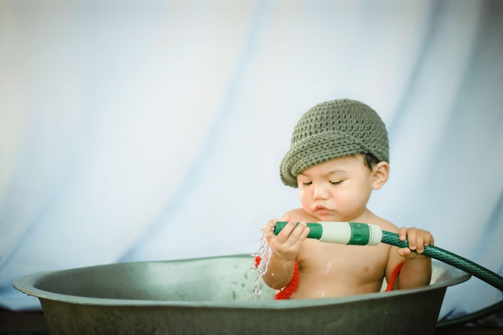 Little boy wearing a crochet cap sitting in a big tub holding an open hose - Baby Names That Mean Curious - Baby Journey