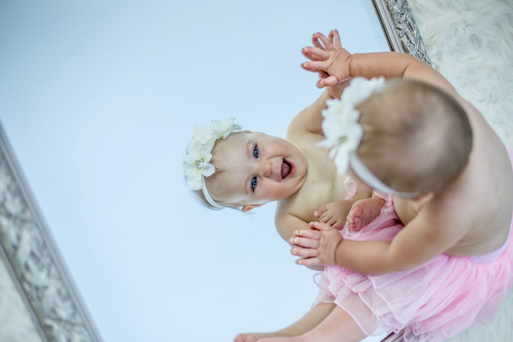 A happy smiling baby girl dressed in pink skirt looking at a large mirror - Catholic baby names - Baby Journey