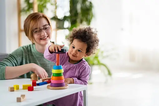 A nanny watching as a toddler plays with a stacking toy - Types of child care - Baby Journey