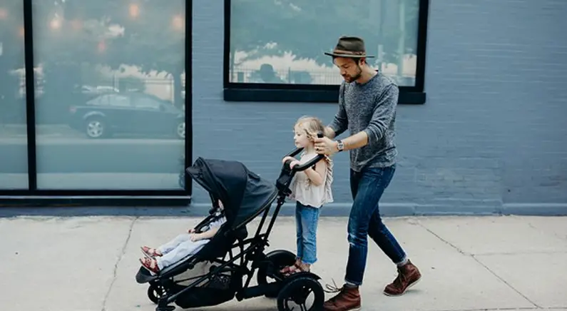 A father pushing a baby stroller with a young girl riding a buggy board - Best Stroller Boards - Baby Journey