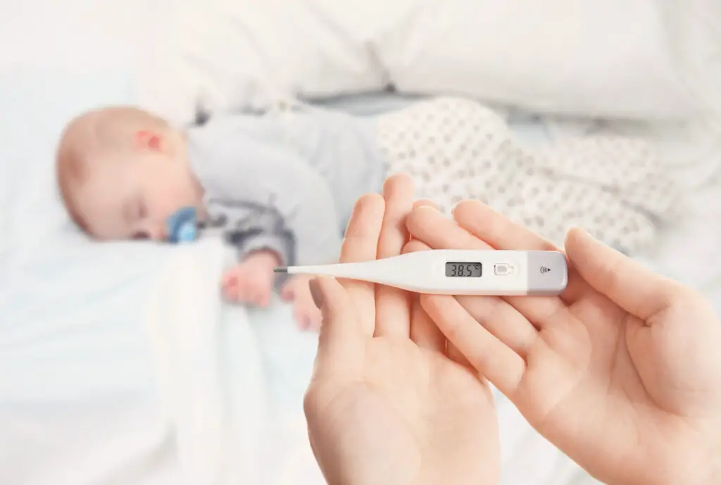  Hands holding a thermometer saying 38.5 degrees Celsius than normal baby temperature while a baby sleeps on the bed - Baby Fever in Infants, High Fever in Kids - babyjourney.net
